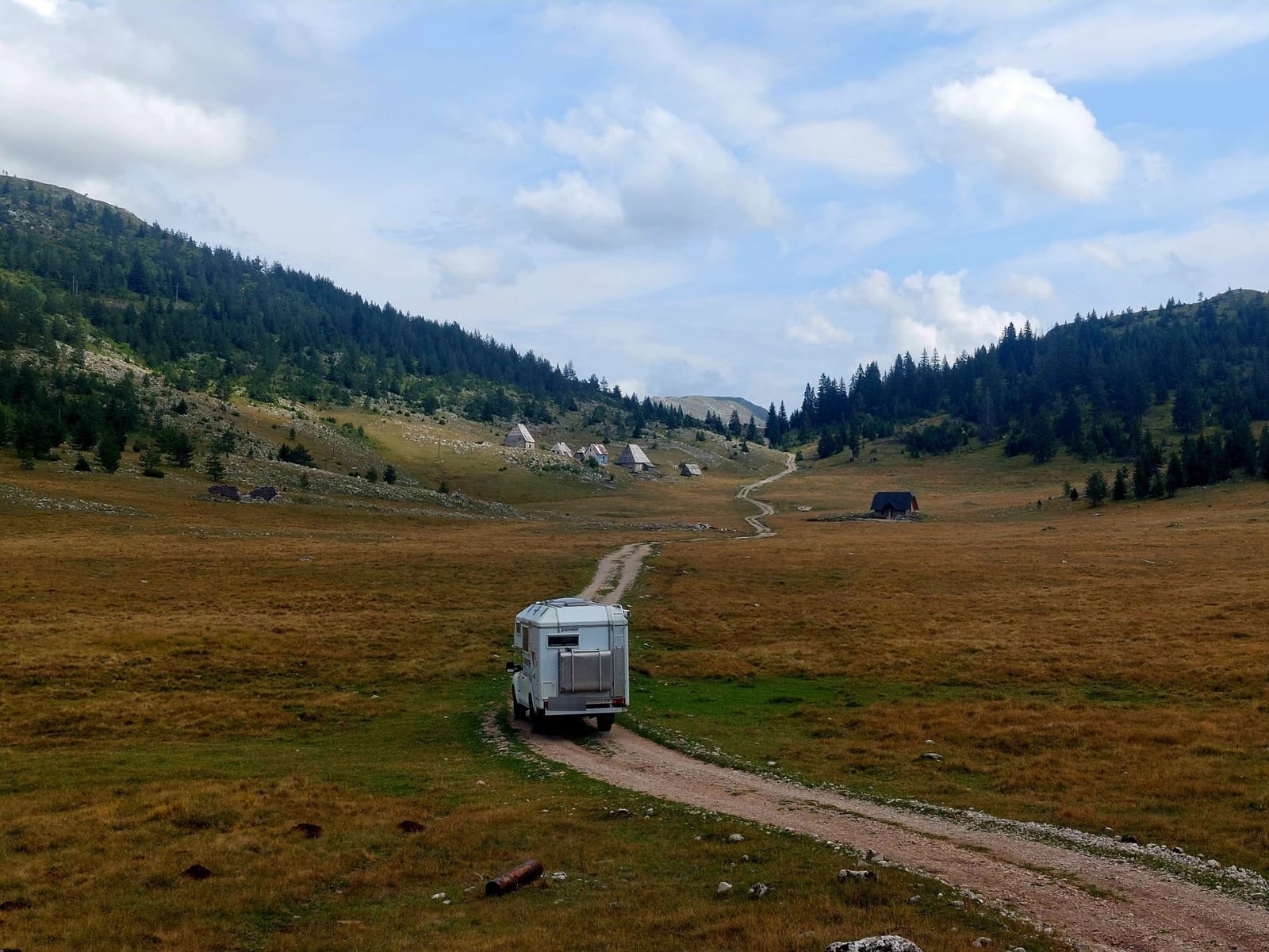 Rancherita por una pista de tierra que cruza un valle de pastos con cabañas de madera y bosque de coníferas